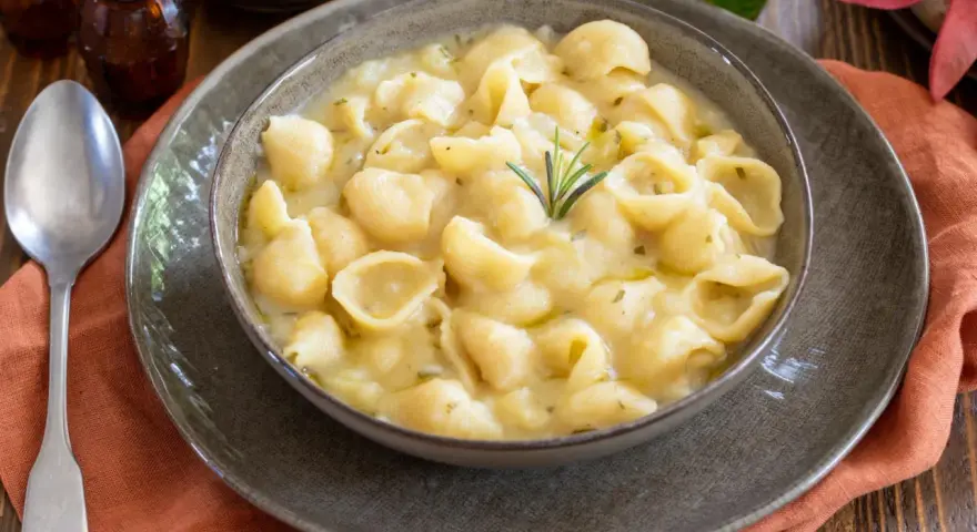 Una ciotola di minestra di patate cremosa con pasta a conchiglia, guarnita con rosmarino. Un piatto confortante e saporito.