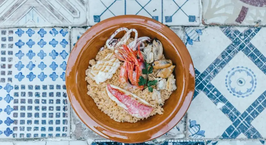 Un invitante cous cous di pesce con gamberi, calamari, cozze e pesce rosso, guarnito con prezzemolo, servito in una ciotola di terracotta su un piatto di maioliche.