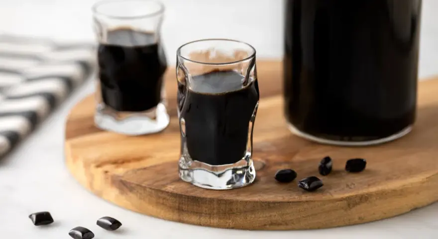 Due bicchierini e una bottiglia di liquore alla liquirizia scuro, con caramelle di liquirizia sparse su un tagliere di legno.