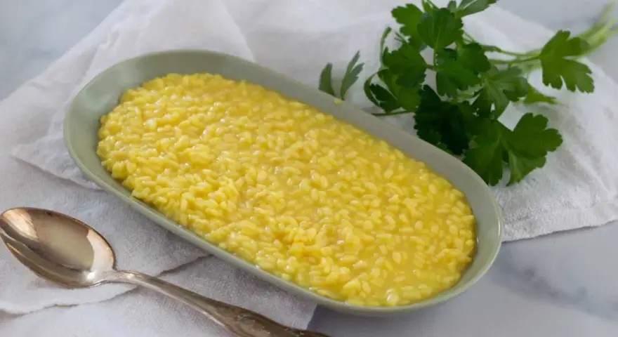 Un cremoso risotto allo zafferano in un piatto ovale, con un cucchiaio d'argento e un rametto di prezzemolo fresco.