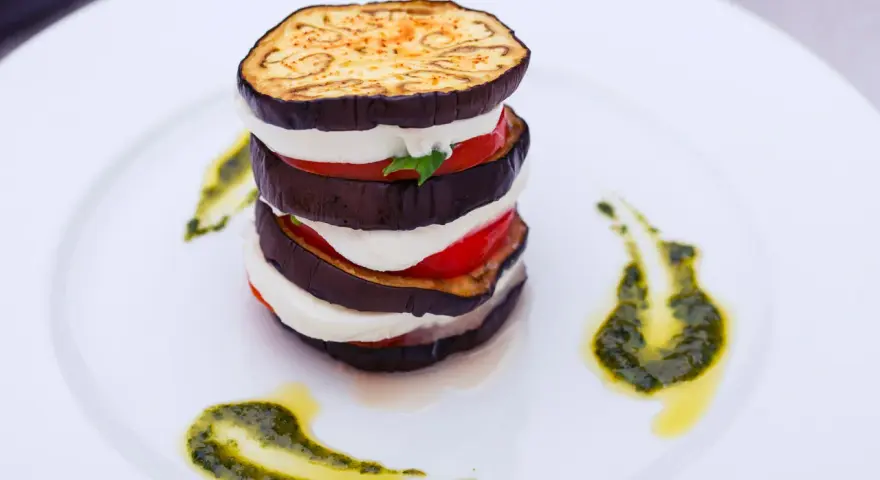 Parmigiana di melanzane a strati, con mozzarella, pomodoro e basilico, guarnita con pesto su piatto bianco.