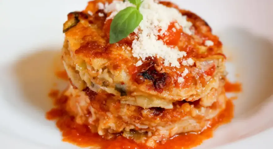 Parmigiana di alici, un piatto stratificato con salsa di pomodoro, formaggio e basilico fresco, servito su un piatto bianco.