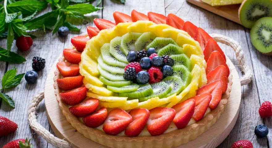 Una colorata crostata di frutta, con fragole, kiwi, ananas e frutti di bosco, su un tavolo di legno rustico.