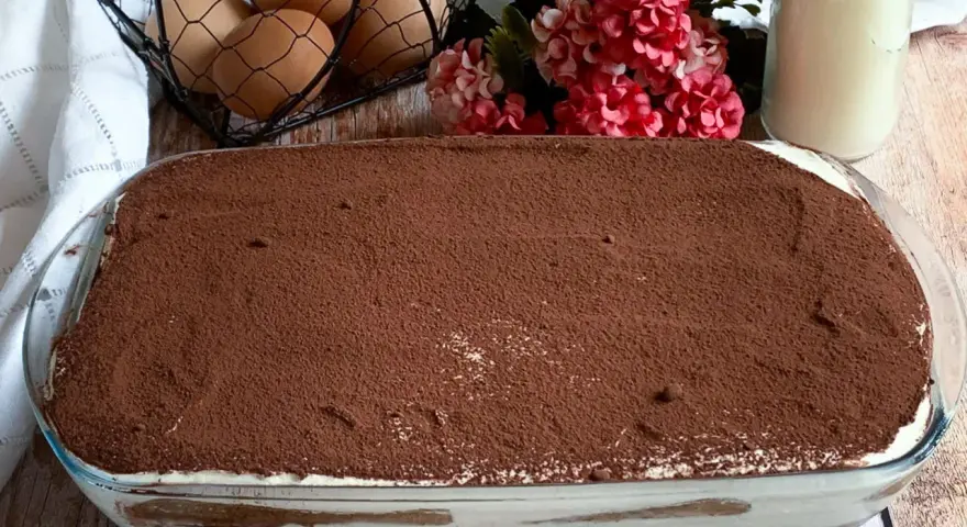 Un invitante tiramisù con panna, cosparso di cacao, pronto per essere gustato. Ingredienti freschi come uova e latte completano la scena.