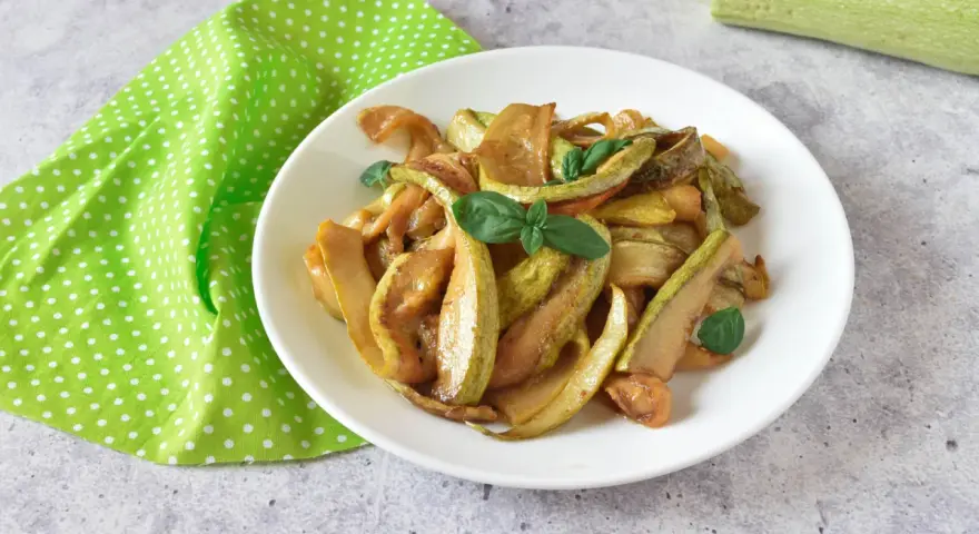 Zucchine in agrodolce, un piatto colorato e saporito, guarnito con basilico fresco su un piatto bianco, accanto a un tovagliolo a pois verdi.
