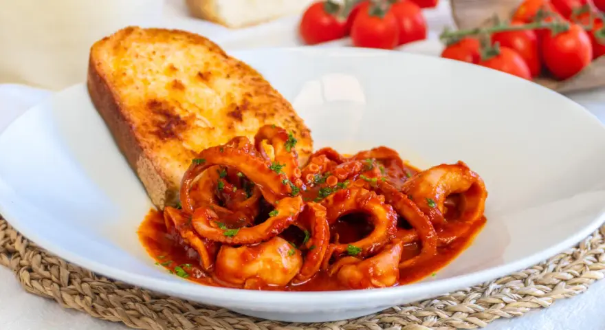 Un piatto invitante di moscardini in umido, con tentacoli succulenti in salsa rossa, accompagnati da una fetta di pane tostato dorato.