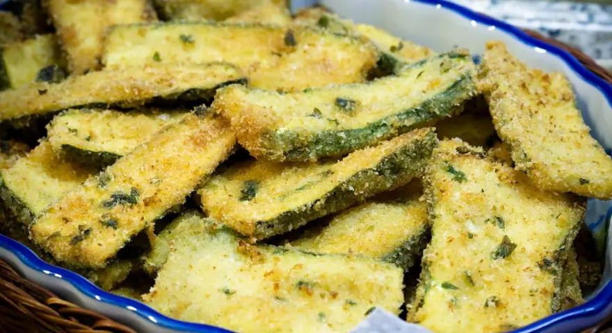 Fette di zucchine gratinate in padella, dorate e croccanti, servite in una ciotola blu bordata di bianco.