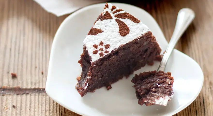 Una fetta di torta caprese ricetta originale napoletana, cosparsa di zucchero a velo e cacao, su un piattino bianco a forma di cuore.