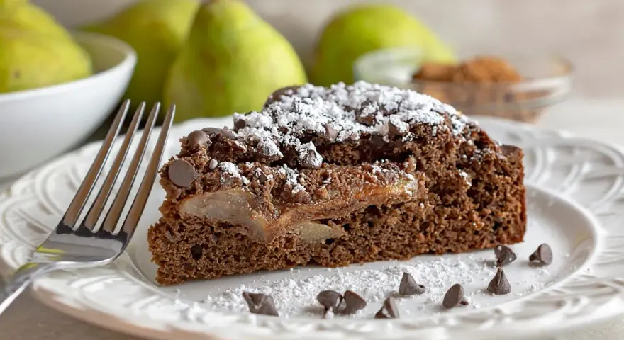 Una fetta di torta pere e cioccolato, cosparsa di zucchero a velo e gocce di cioccolato, con pere caramellate all'interno.