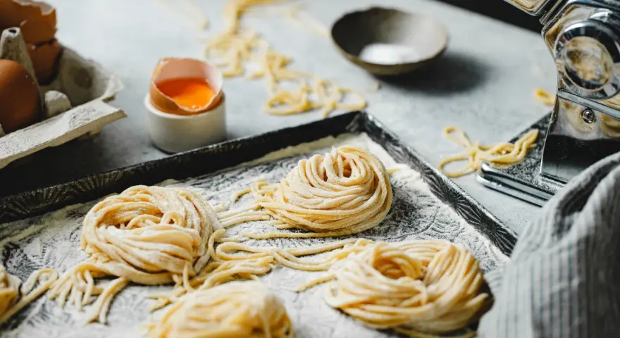 Nidi di pasta fatta in casa su un vassoio infarinato, con uova e una macchina per la pasta sullo sfondo.