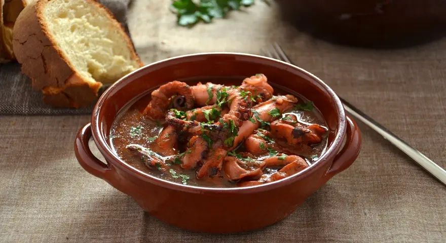 Un saporito polpo alla pignata, cotto lentamente in umido, servito in una ciotola di terracotta con pane fresco a lato.
