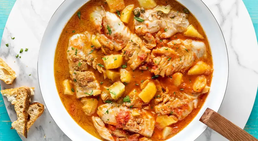 Un saporito merluzzo in umido con patate e pomodoro, guarnito con prezzemolo fresco e accompagnato da pane tostato.