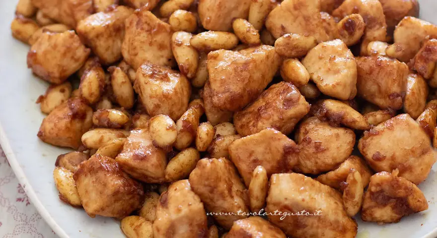 Close-up of bite-sized chicken pieces and almonds coated in a glossy brown sauce on a white plate.
