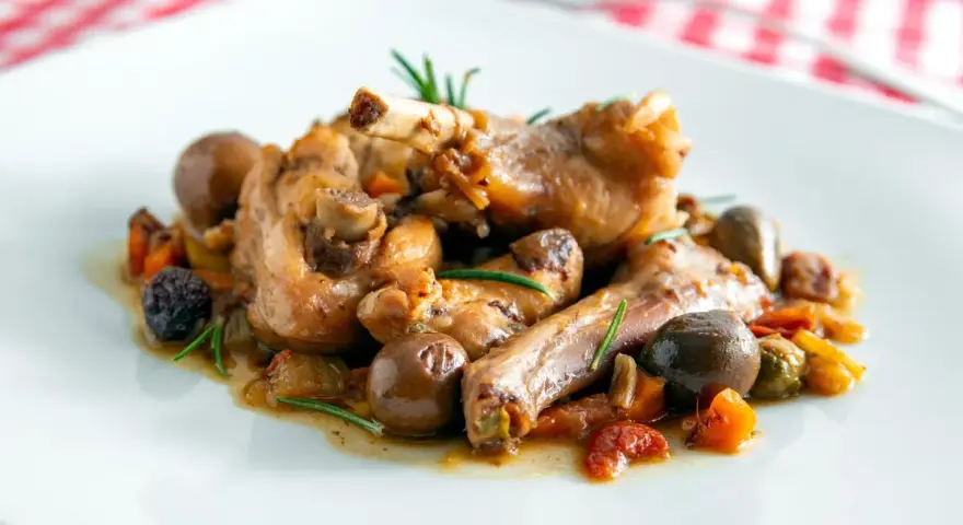 Coniglio alla cacciatora ricetta della nonna: succulenti pezzi di coniglio con olive, pomodorini e rosmarino su un piatto bianco.