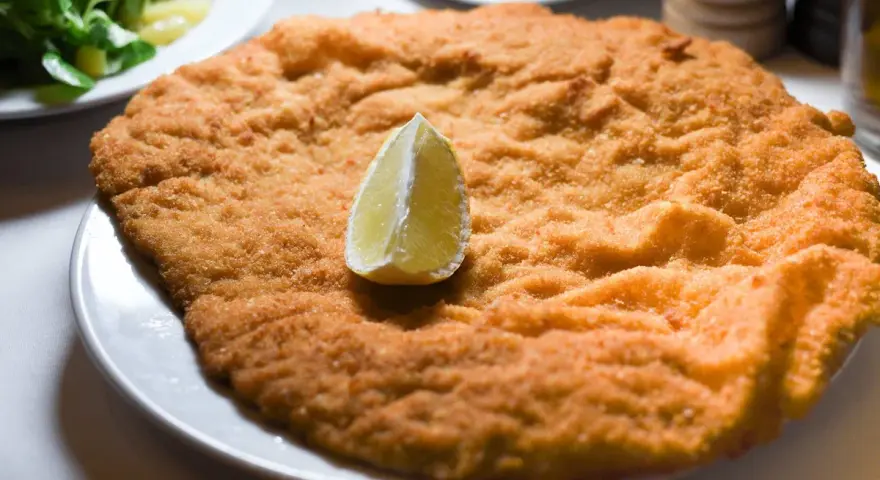 Una cotoletta dorata, grande come un'orecchia di elefante, servita con una fetta di limone e un'insalata fresca.