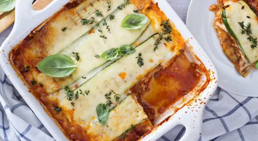 Lasagne di zucchine al forno, dorate e filanti, guarnite con basilico e timo. Una porzione è già servita su un piatto bianco.