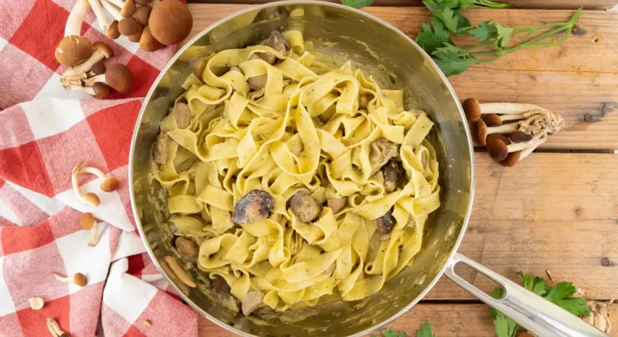 Tagliatelle ai funghi cremose in padella, con funghi freschi e prezzemolo su fondo di legno.