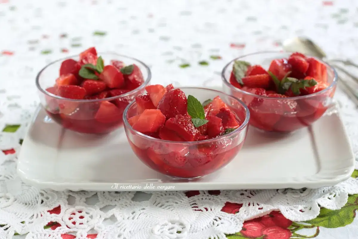 Tre coppette di macedonia di frutta con fragole fresche e foglie di menta, servite su un vassoio bianco.