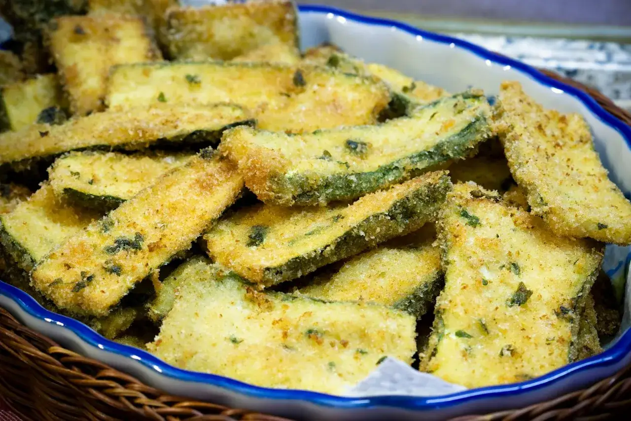 Fette di zucchine gratinate in padella, dorate e croccanti, servite in una ciotola blu bordata di bianco, pronte per essere gustate.