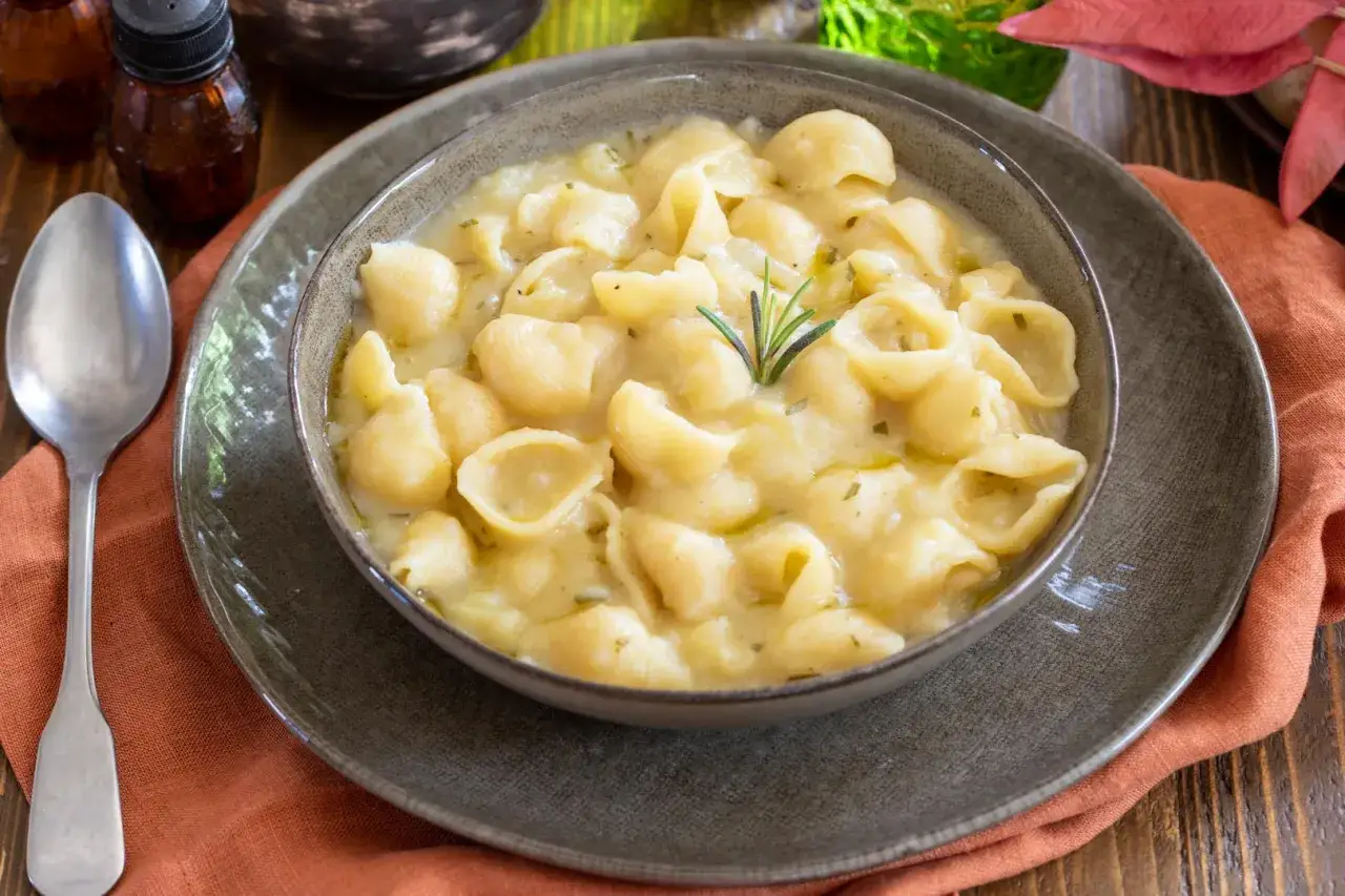 Una ciotola di minestra di patate cremosa con pasta a conchiglia, guarnita con rosmarino. Un piatto confortante e saporito.