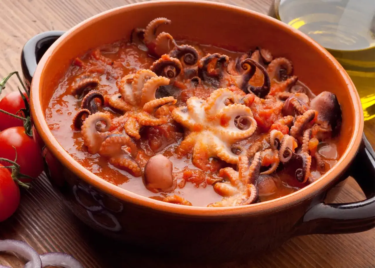 Un saporito polpo alla luciana in un piatto di terracotta, con pomodori freschi e cipolla rossa a lato.