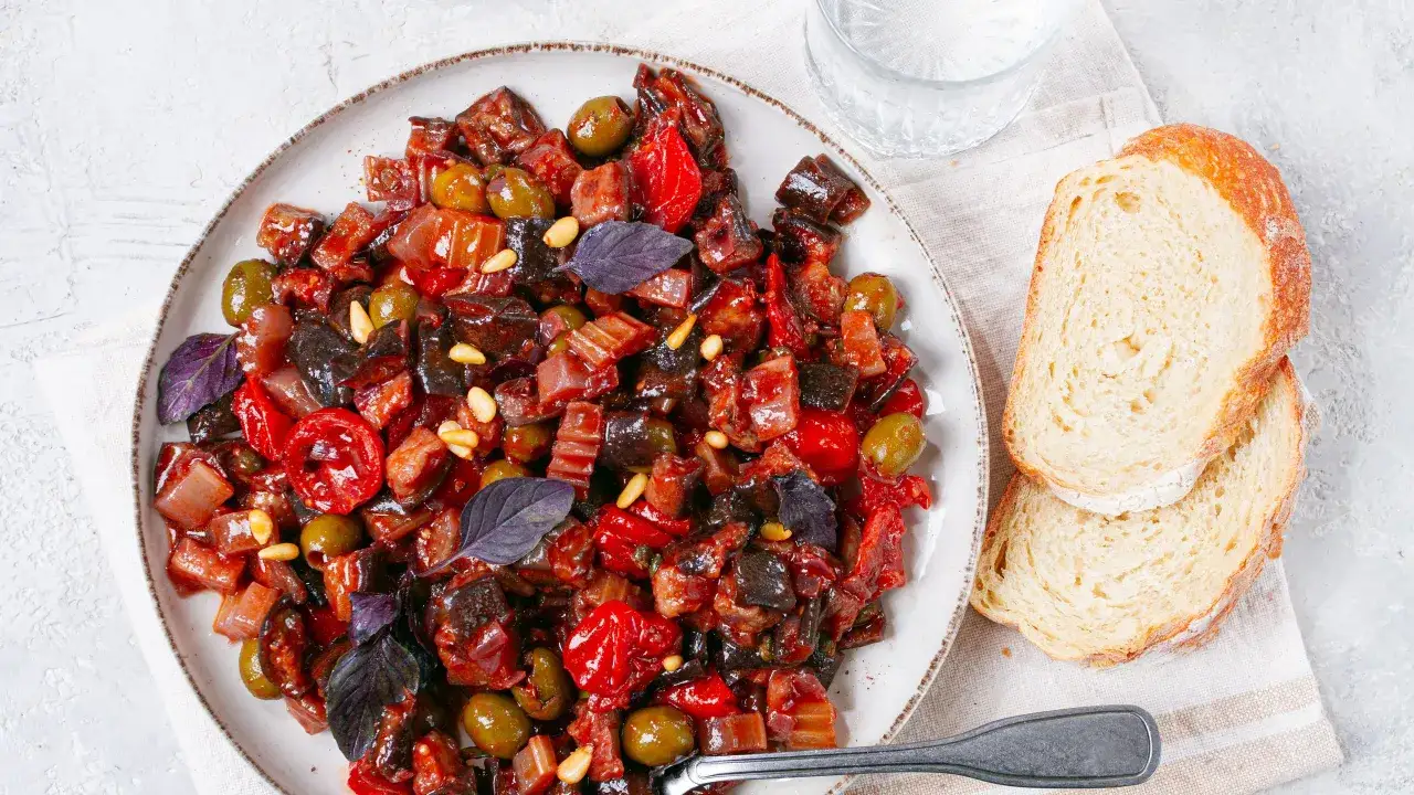 Un piatto invitante di caponata di verdure, ricca di melanzane, pomodori, olive e pinoli, guarnita con basilico viola. Accompagnata da fette di pane rustico.