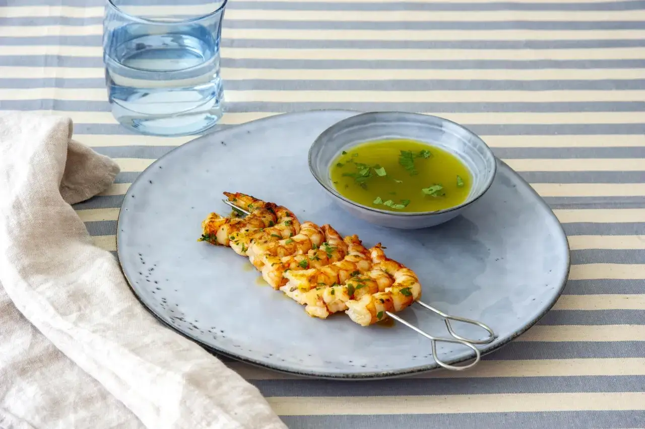 Gamberoni alla griglia dorati e succulenti su uno spiedino, accompagnati da una ciotolina di salsa verde e un bicchiere d'acqua.