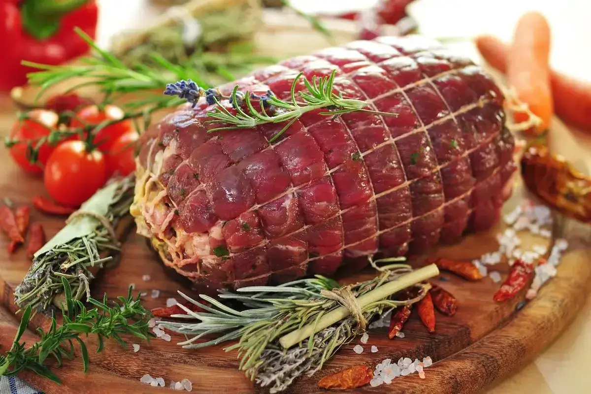 Un succulento arrosto di manzo pronto per essere cucinato, circondato da erbe aromatiche, pomodorini, peperoncini e carote.