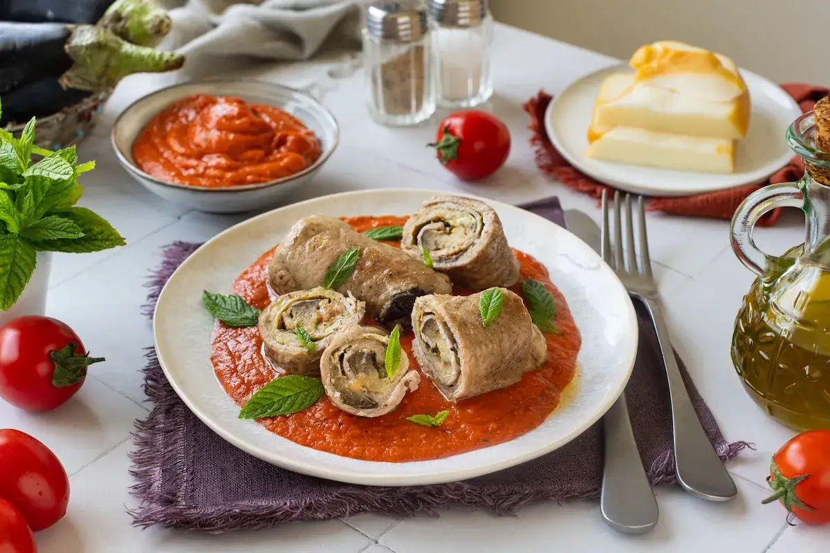 Involtini di melanzane alla siciliana su un letto di salsa rossa, guarniti con menta fresca. Accanto, pomodori, formaggio e olio d'oliva.