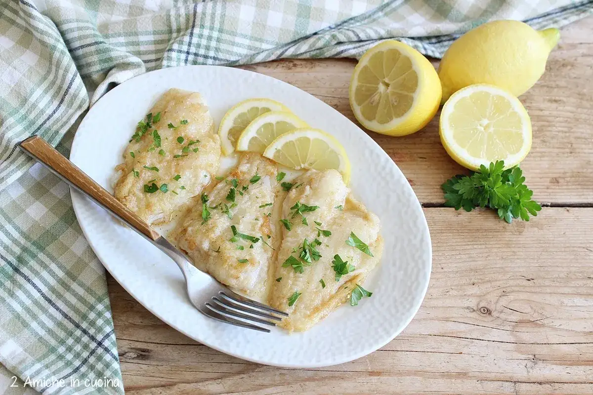 Sogliola alla mugnaia servita su un piatto bianco, guarnita con prezzemolo e fette di limone. Accanto, limoni freschi e un rametto di prezzemolo.