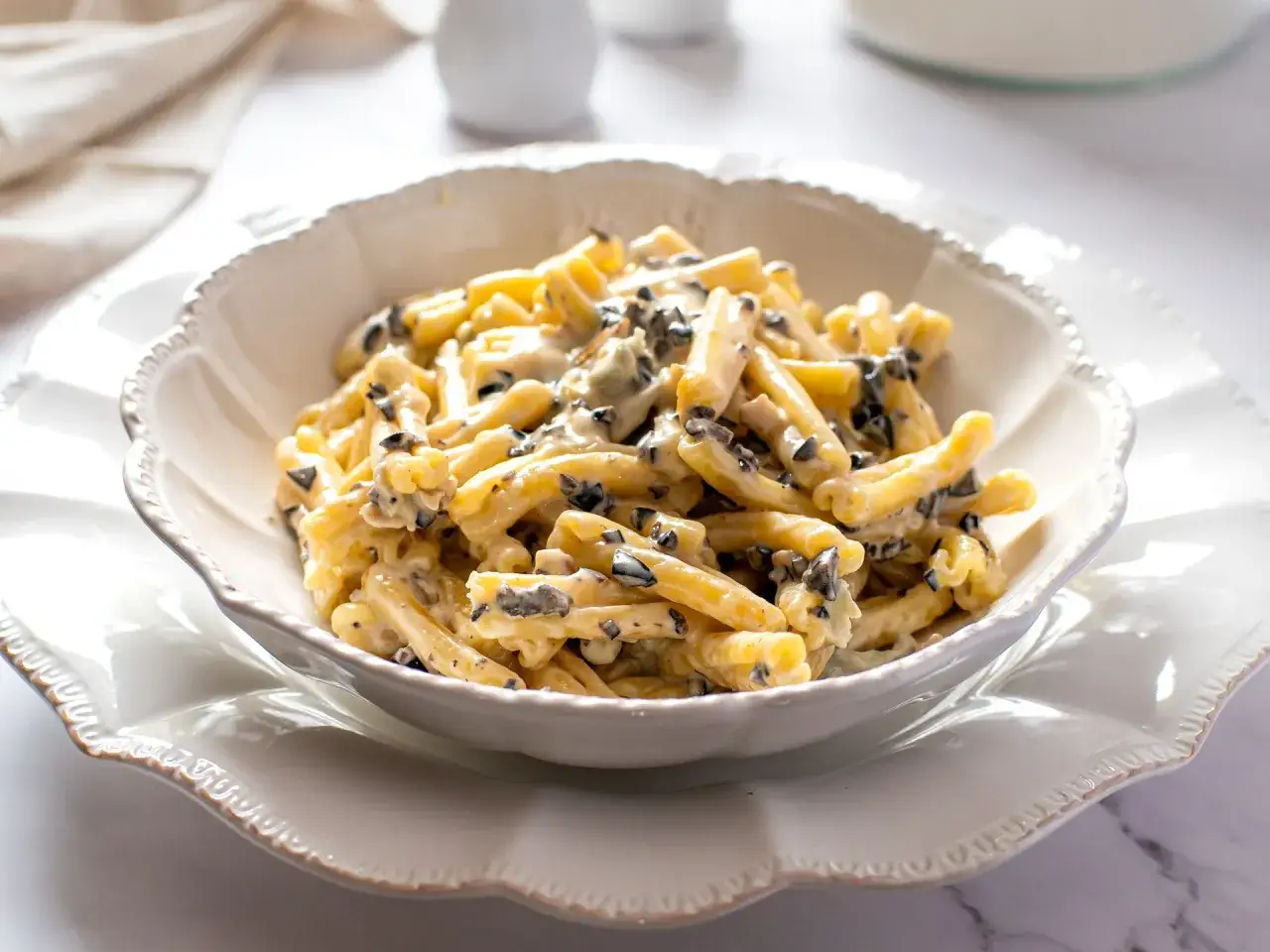 Un piatto di pasta alla cenere, condita con una cremosa salsa e pezzetti scuri, servita in una ciotola bianca decorata.