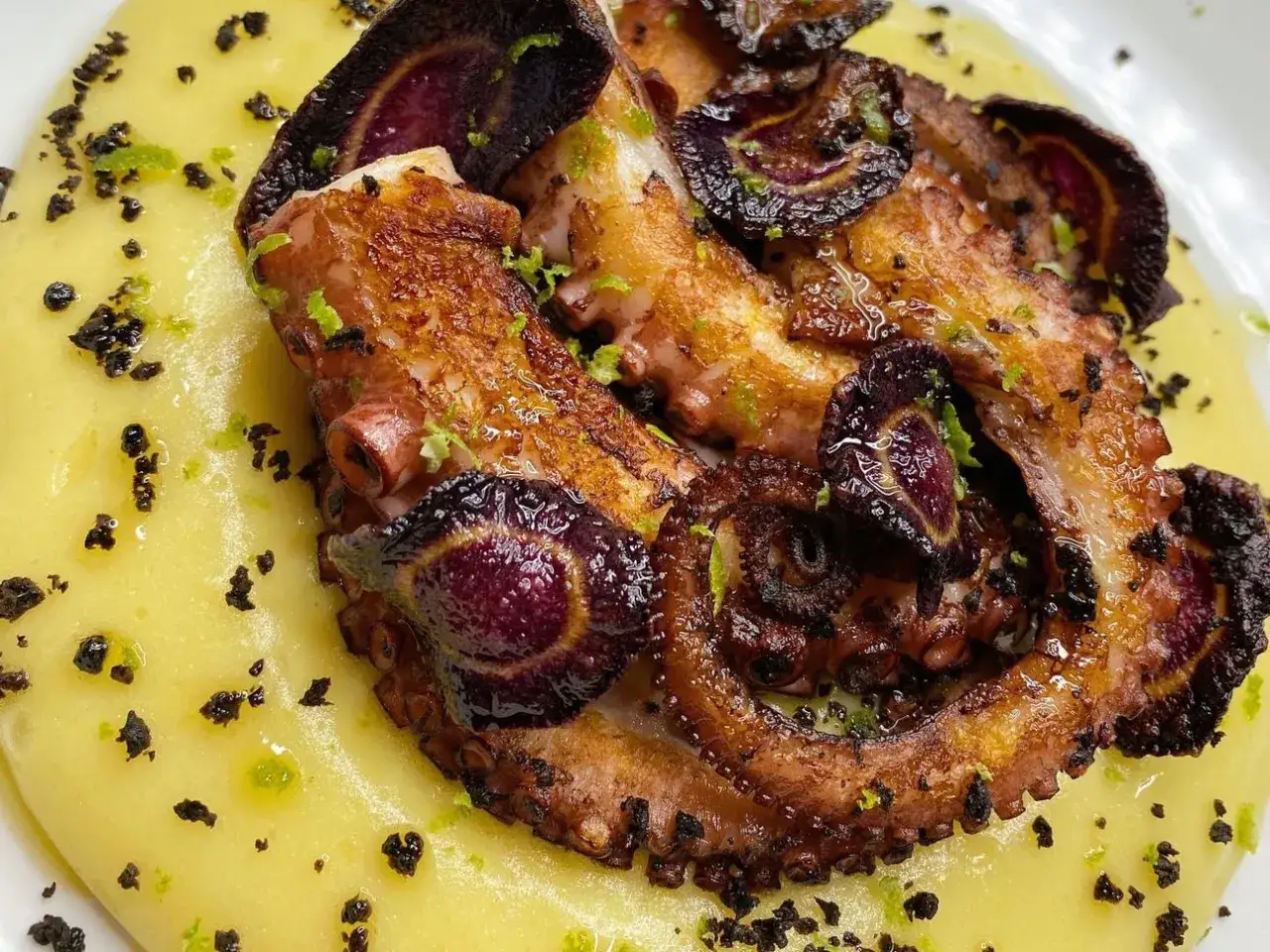 Polpo alla gallega succulento su crema gialla, guarnito con anelli di carota viola e scorza di lime.