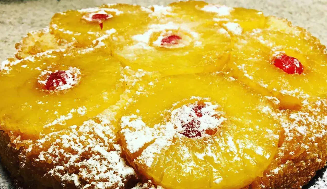 Torta rovesciata all'ananas, con fette di ananas caramellato e ciliegie rosse, spolverata di zucchero a velo.