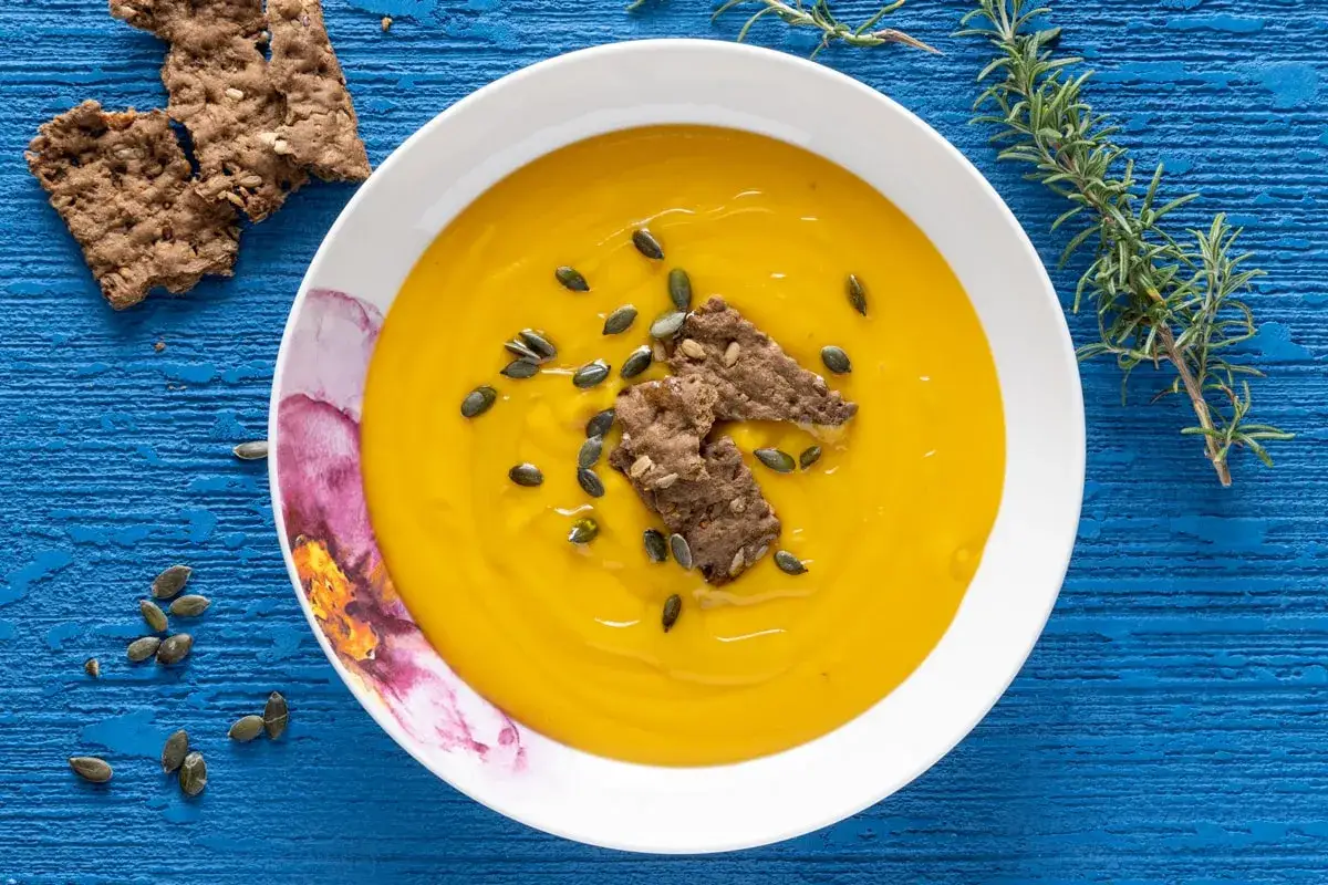 Vellutata di zucca cremosa, guarnita con semi di zucca e crostini integrali, su un tavolo blu. Un rametto di rosmarino completa la composizione.