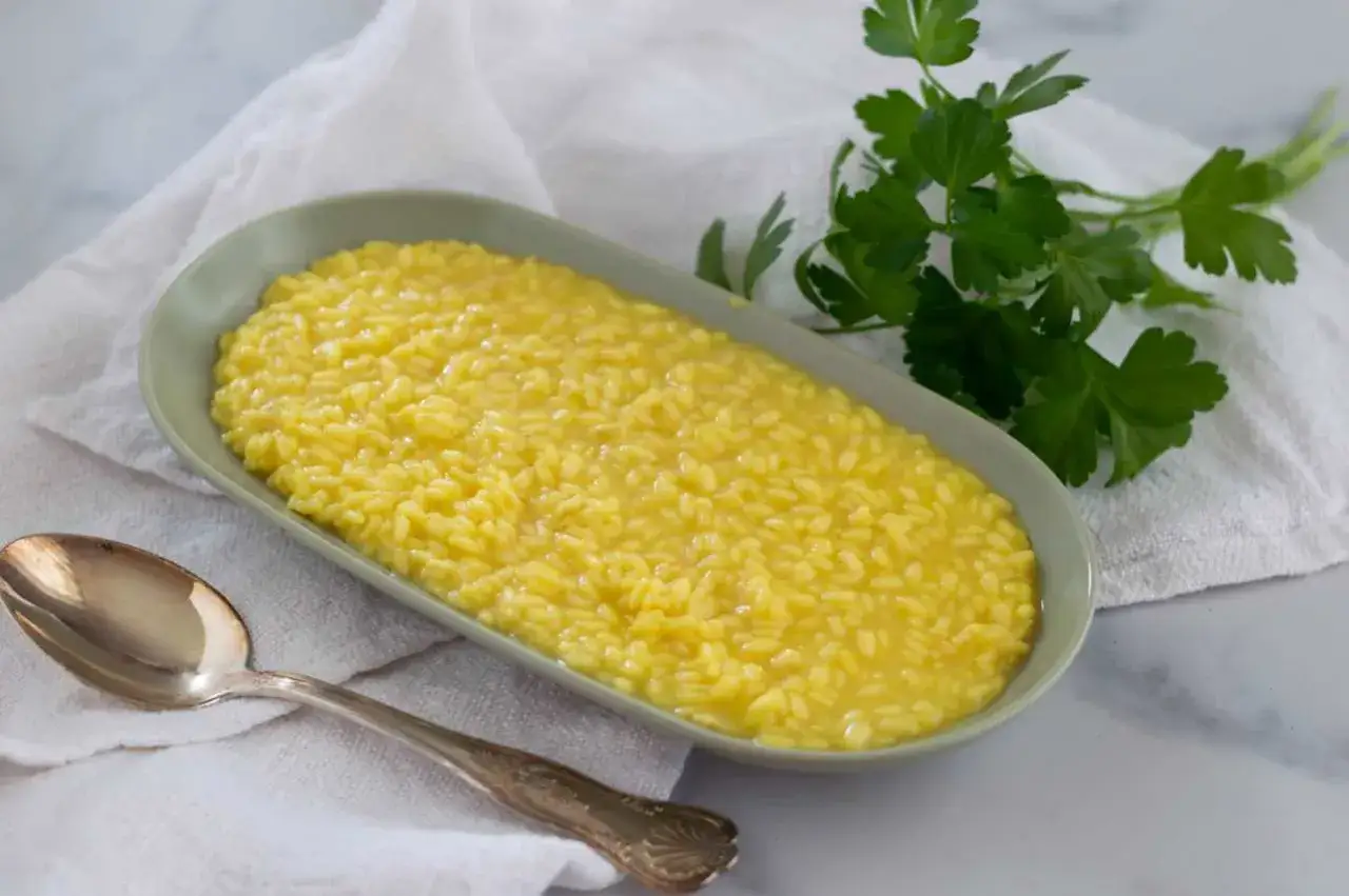 Un cremoso risotto allo zafferano in un piatto ovale, con un cucchiaio d'argento e un rametto di prezzemolo fresco.