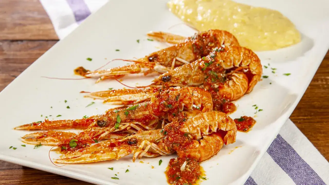 Scampi alla busara succulenti su un piatto bianco, conditi con salsa rossa e prezzemolo, accompagnati da una porzione di polenta.
