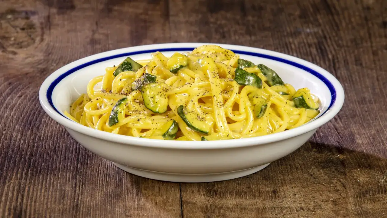 Spaghetti cremosi con zucchine e pepe nero, una deliziosa carbonara di zucchine su un piatto bianco con bordo blu, su un tavolo di legno rustico.
