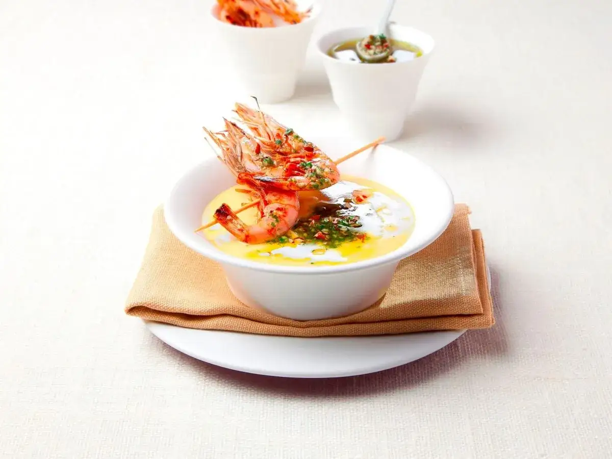 Deliziosa bisque di gamberi servita in una ciotola bianca, guarnita con gamberi arrostiti su uno spiedino e un filo d'olio aromatico.
