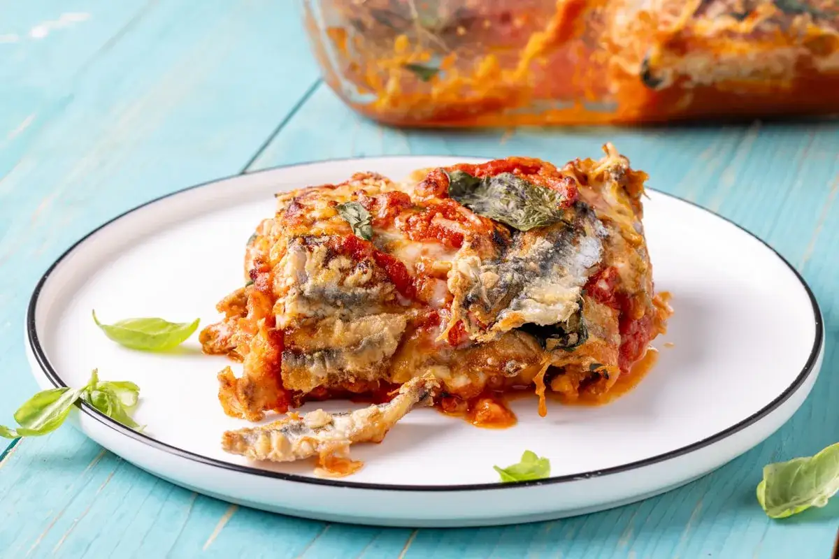 Una porzione di parmigiana di alici, ricca di pomodoro e basilico, servita su un piatto bianco con sfondo azzurro.