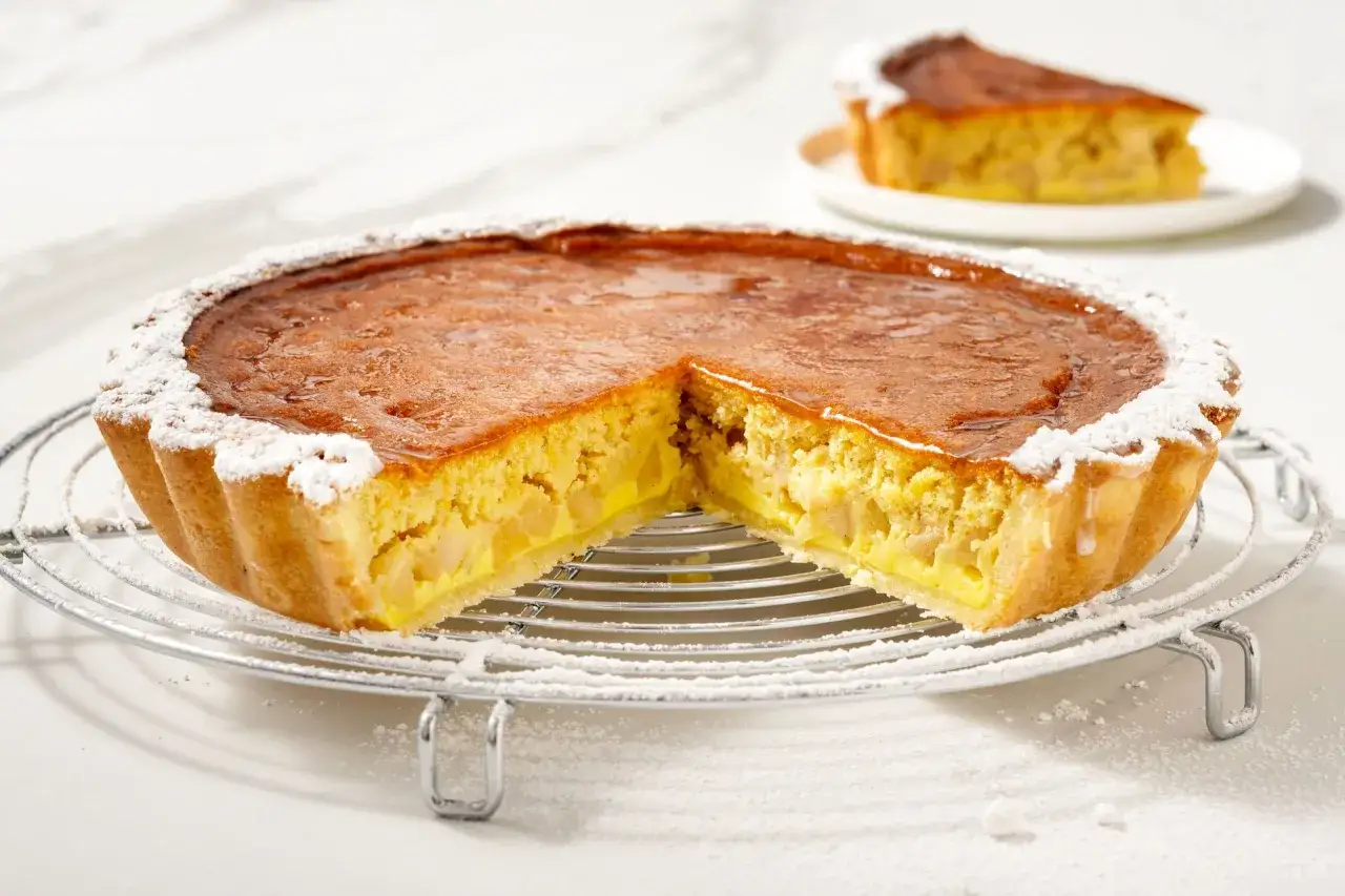 Una deliziosa torta di mele con crema pasticcera, cosparsa di zucchero a velo, su una gratella. Una fetta &egrave; gi&agrave; servita su un piattino.