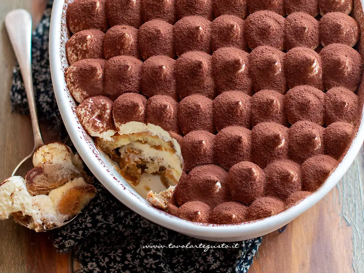 Un invitante tiramis&ugrave; ricetta originale, cosparso di cacao, in una teglia bianca. Un cucchiaio ne serve una porzione cremosa.
