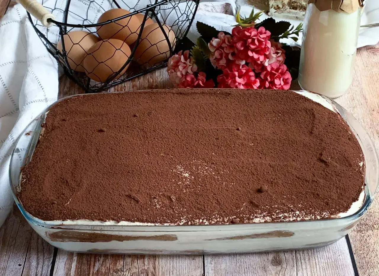 Un invitante tiramis&ugrave; con panna, cosparso di cacao, pronto per essere gustato. Ingredienti freschi come uova e fiori decorano la scena.