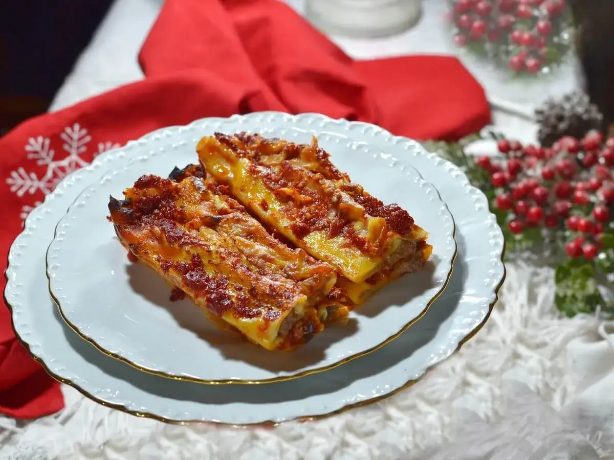 Due cannelloni ripieni di carne macinata e mozzarella, cotti al forno e serviti su un piatto bianco con decorazioni natalizie sullo sfondo.