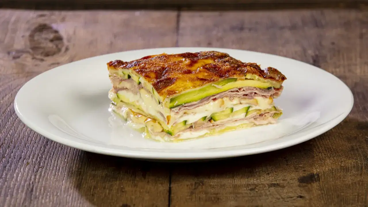 Una fetta di parmigiana di zucchine bianca senza besciamella, con strati di zucchine, prosciutto e formaggio filante, gratinata in forno.