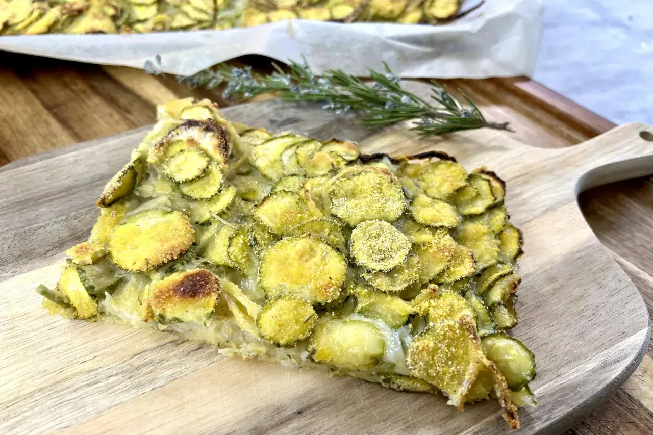 Una fetta di scarpaccia di zucchine dorata e croccante, con zucchine affettate e panatura, su un tagliere di legno.