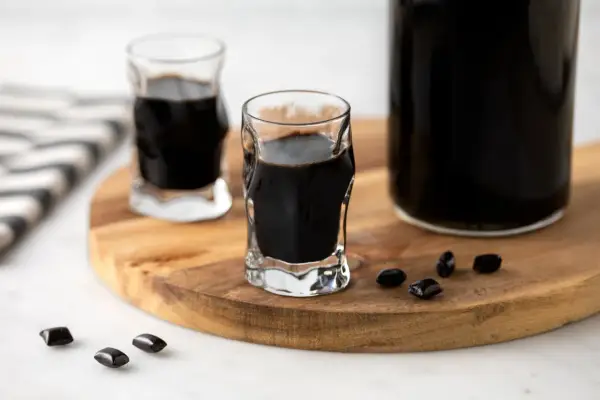 Due bicchierini e una bottiglia di liquore alla liquirizia scuro, con caramelle di liquirizia sparse su un tagliere di legno.