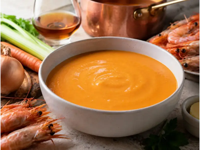 Una ciotola di cremosa bisque di gamberi, con gamberi freschi, carote, sedano e cipolla sullo sfondo.