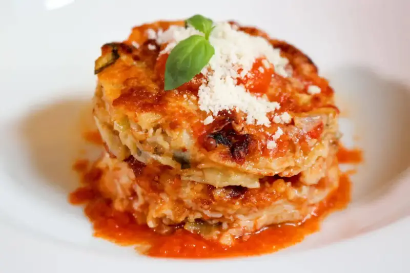 Parmigiana di alici, un piatto stratificato con salsa di pomodoro, formaggio e basilico fresco, servito su un piatto bianco.