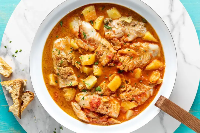 Un saporito merluzzo in umido con patate e pomodoro, guarnito con prezzemolo fresco e accompagnato da pane tostato.